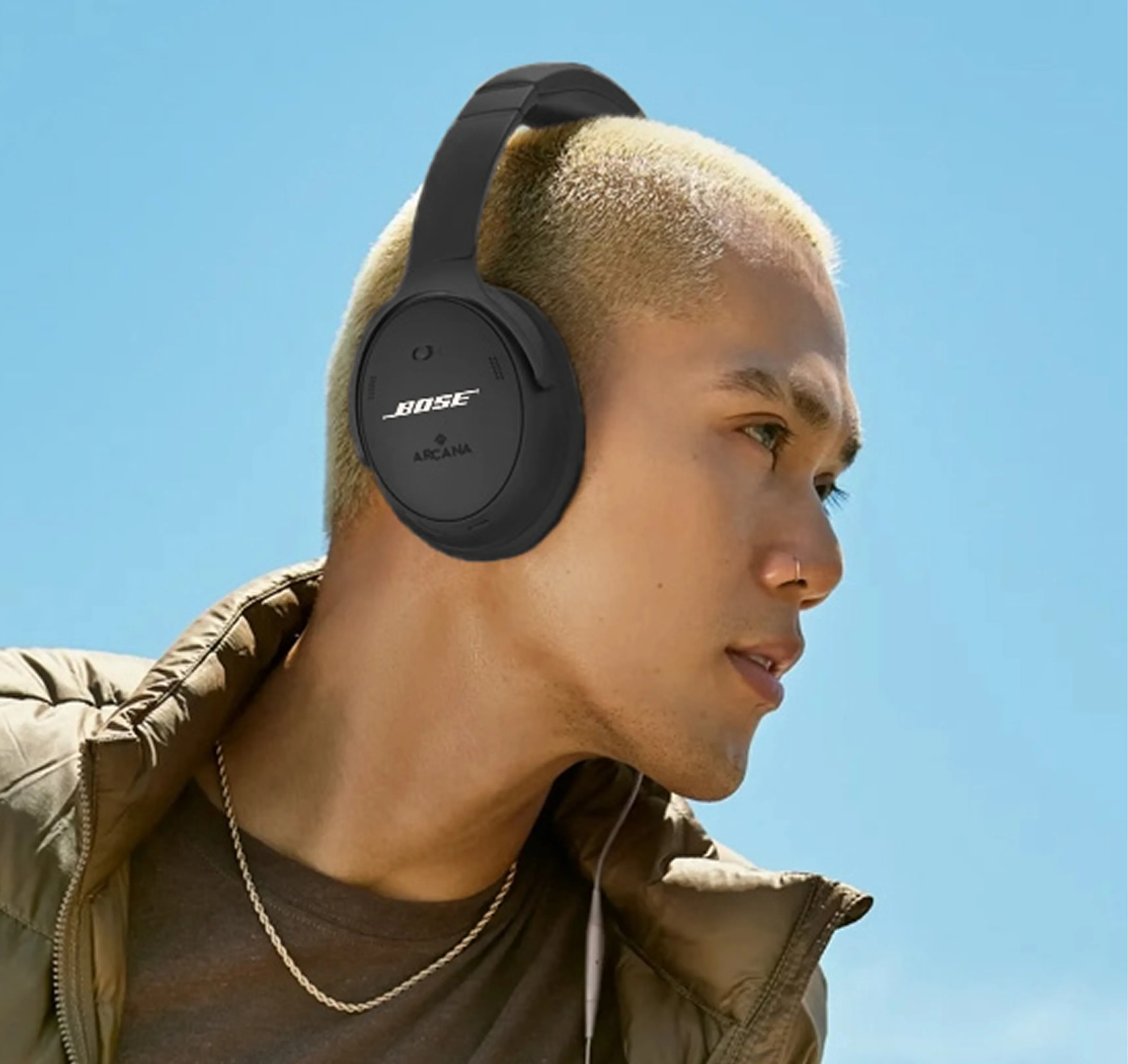 Young man with short blond hair wearing black Bose over-ear headphones and a chain necklace against a clear blue sky.