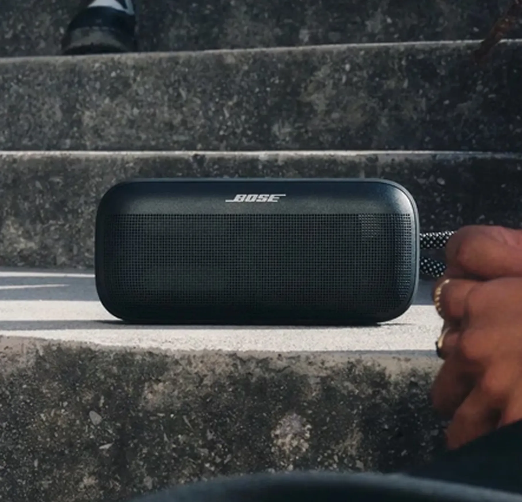 Black Bose portable speaker placed on concrete steps with a person's hand holding its strap.