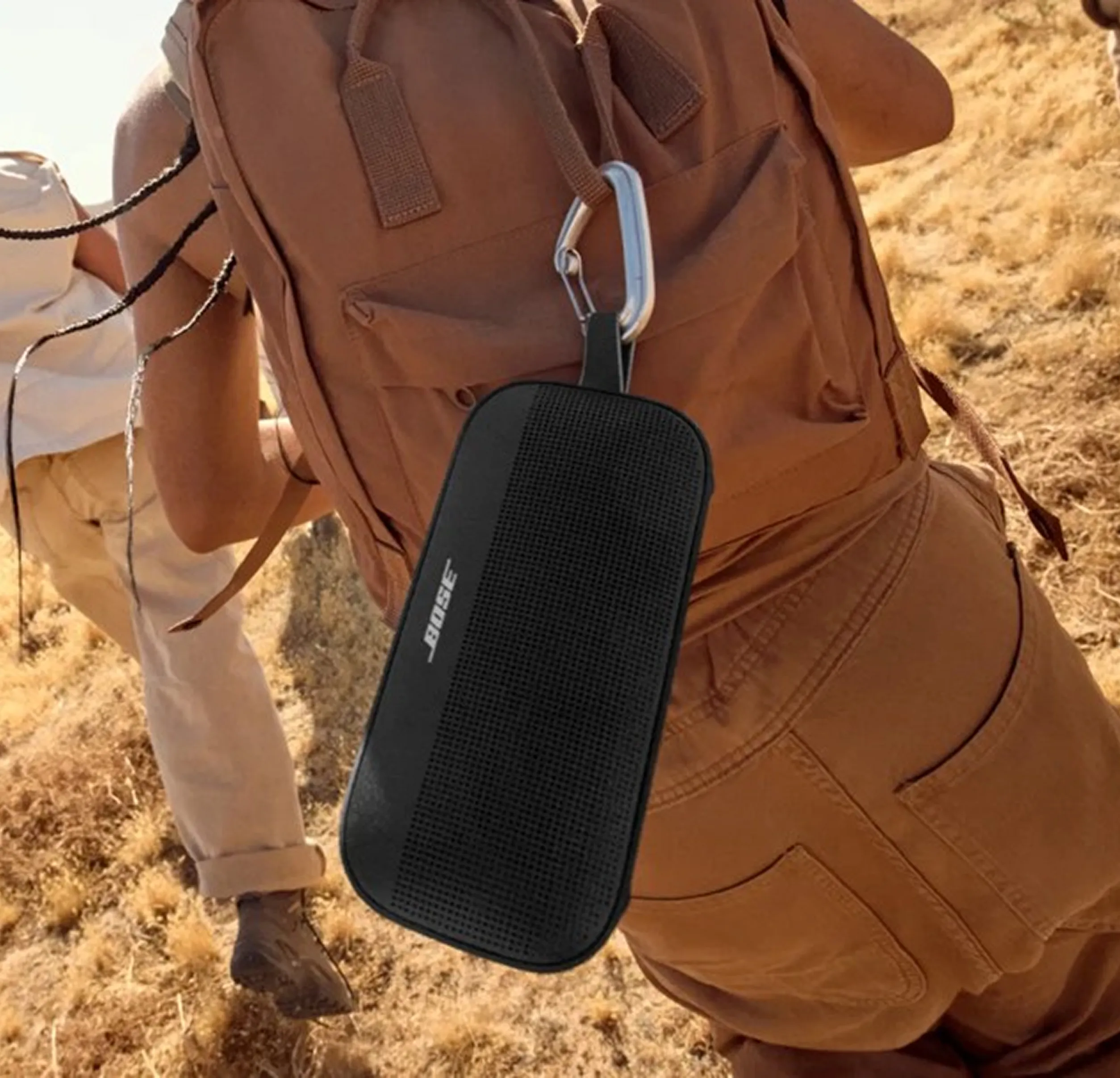 Black Bose portable speaker clipped to a brown backpack worn by a person hiking on dry grass.